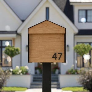Peut inclure: Une bo&icirc;te aux lettres rectangulaire avec un toit en pignon, pr&eacute;sentant une finition en grain de bois brun clair et le num&eacute;ro 47 imprim&eacute; en noir sur le devant. La bo&icirc;te aux lettres est mont&eacute;e sur un poteau m&eacute;tallique noir, fin et haut.