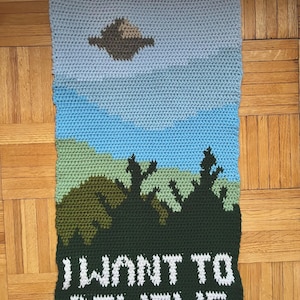 Könnte beinhalten: Gehäkeltes Wandbild mit einer Landschaft mit einem UFO und dem Text "I WANT TO BELIEVE". Das Kunstwerk verwendet Blau-, Grün- und Brauntöne, um einen Himmel, Bäume und eine fliegende Untertasse darzustellen. Der Text ist weiß.