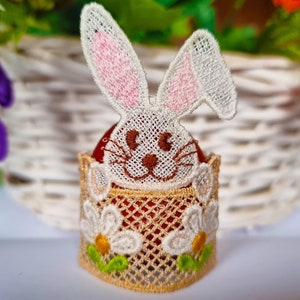 May include: A white crocheted bunny rabbit with pink ears sits on top of a brown egg. The egg is decorated with a white crocheted lace design and green leaves.