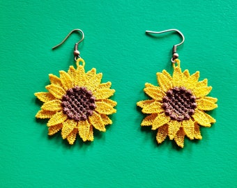 Puede incluir: Un par de pendientes de girasol. Cada pendiente presenta un girasol amarillo, similar al encaje, con un centro marrón. Los pendientes tienen ganchos de color bronce y están sobre un fondo verde. Los pendientes son un accesorio floral.