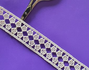 May include: White lace trim with a delicate pattern of small circles and diamonds. The trim is lying on a purple surface with a pair of silver scissors.