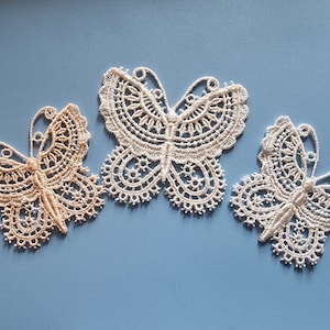 May include: Three white lace butterfly embellishments on a light blue background. Two of the butterflies are white and one is a light beige colour.