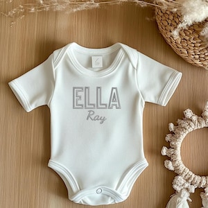 Op de afbeelding: Witte baby romper met grijze tekst die "ELLA Ray" luidt.