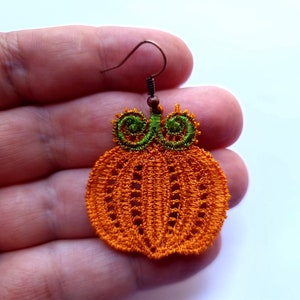 May include: Orange and green pumpkin-shaped earrings made of crocheted thread. The earrings are hanging from a brown metal hook.