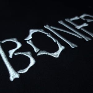 May include: A black t-shirt with the word "BONES" embroidered in white, the letters are stylized to look like bones.