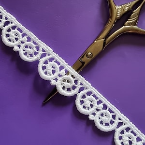 Könnte beinhalten: Weiße Spitzenborte mit einer silbernen Schere auf einem violetten Hintergrund.