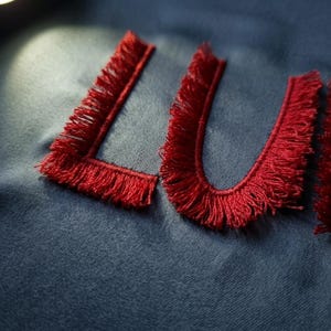 Puede incluir: Una superficie de tela azul oscuro con las letras L y U bordadas en hilo rojo intenso. Las letras están delineadas con un fleco denso y texturizado de hilo de bordar rojo, creando un efecto elevado y decorativo sobre el material liso.