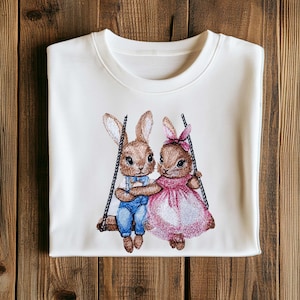 Könnte beinhalten: Weißer Pullover mit gesticktem Bild von zwei braunen Kaninchen auf einer Schaukel. Das Kaninchen links trägt eine blaue Latzhose und eine Fliege. Das Kaninchen rechts trägt ein rosa Kleid mit Schleife.