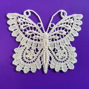 May include: A white lace butterfly applique with intricate details. The butterfly is on a purple background.