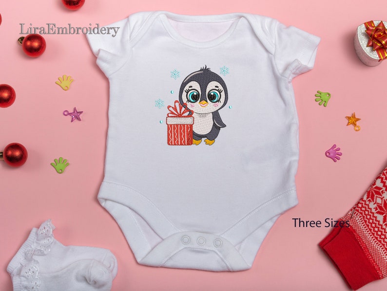 May include: White baby onesie with an embroidered design of a penguin holding a red and white gift box. The penguin is wearing a black hat and has blue eyes. The design is surrounded by snowflakes.