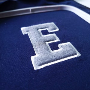 May include: Close-up of navy blue fabric with a grey, embroidered letter "E". The letter has a white border and is stitched onto the fabric. The fabric is held in a white embroidery hoop.