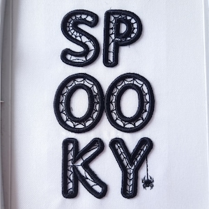 Könnte beinhalten: Schwarze gestickte Buchstaben, die "SPOOKY" buchstabieren, mit einer kleinen schwarzen Spinne, die am "Y" hängt.