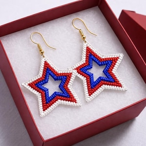May include: A pair of star-shaped earrings in a red gift box. The earrings feature a white outline, with red and blue inner star details. The earrings have gold-colored hooks.