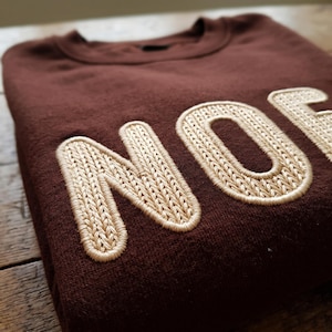 Puede incluir: Una sudadera de cuello redondo en color marrón oscuro con grandes letras bordadas en color crema que forman la palabra NOEL. El bordado tiene una textura en relieve similar al tejido de punto y las letras están delineadas con una puntada gruesa.