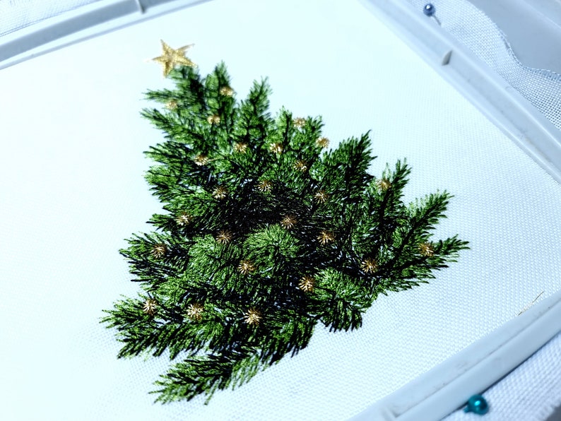 May include: Embroidered Christmas tree design on white fabric with gold accents. The tree is green and has a star on top.