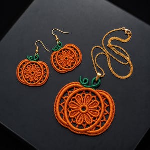 Può includere: Un set di gioielli composto da due orecchini a forma di zucca e un ciondolo abbinato su una catenina dorata. I pezzi sono realizzati in filo arancione con un intricato ricamo floreale e presentano piccoli steli in filo verde in cima.
