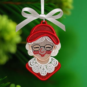 以下が含まれることがあります： 刺繍されたクリスマスオーナメント、ミセス・クロースのデザイン。オーナメントは赤い帽子と襟、白い髪とトリム、黒いメガネが特徴です。白いリボンで吊り下げられ、緑色の背景に白いリボンで結ばれています。