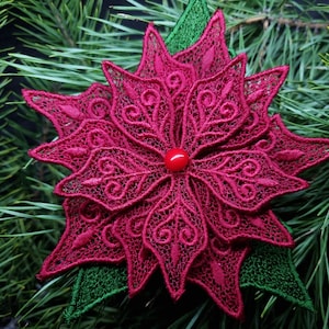 Puede incluir: Un adorno de flor de poinsettia roja hecho de encaje con un acento de hoja verde. El adorno tiene una cuenta roja en el centro.