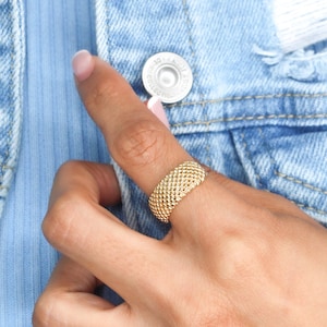 May include: A gold-colored ring with a woven texture is displayed. The ring is worn on a finger, set against a light blue shirt and a denim jacket. The ring's design features a series of small, connected beads, creating a textured surface.