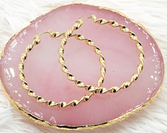 May include: A pair of gold-toned hoop earrings with a twisted design. The earrings are displayed on a pink and gold display stand.