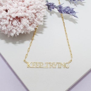 May include: Gold chain necklace with a gold pendant that says "KEEP TRYING".