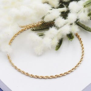 May include: A gold-coloured rope chain necklace is displayed on a white surface, with white fluffy flowers and green leaves. The necklace is a delicate piece of jewellery, suitable for everyday wear.