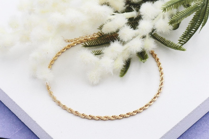 May include: A gold-coloured rope chain necklace is displayed on a white surface, with white fluffy flowers and green leaves. The necklace is a delicate piece of jewellery, suitable for everyday wear.