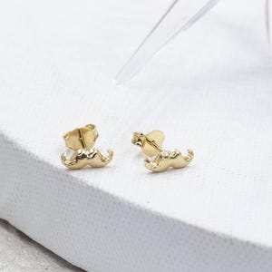 May include: A pair of gold-colored stud earrings shaped like mustaches. The earrings are small and detailed, with a curved design. They are set against a white background.