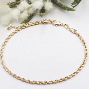 May include: A gold-coloured rope chain bracelet. The bracelet is displayed on a white surface with a blue border, and is accented by white flowers and green leaves in the background. The bracelet has a lobster clasp closure.