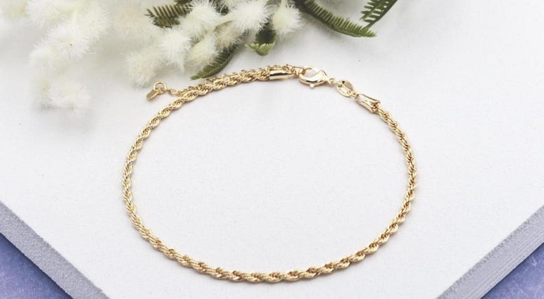 May include: A gold-coloured rope chain bracelet. The bracelet is displayed on a white surface with a blue border, and is accented by white flowers and green leaves in the background. The bracelet has a lobster clasp closure.