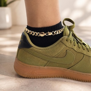 May include: A gold-toned metal curb chain anklet featuring interlocking links and a lobster claw clasp, worn above a black sock and an olive green canvas sneaker with a gum rubber sole and the word AIR embossed on the side heel area.