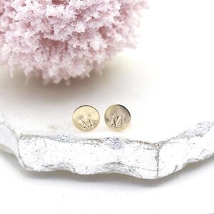 May include: A pair of gold-colored stud earrings, each with the letter "M" engraved on the front. The earrings are round and sit on a white and silver surface, with a pink, fluffy object in the background.