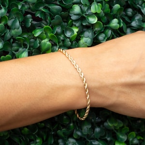 May include: A gold rope chain bracelet on a person's wrist. The bracelet is made of a twisted rope design.