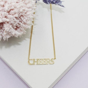 May include: A gold chain necklace with a gold pendant that says "CHEERS".