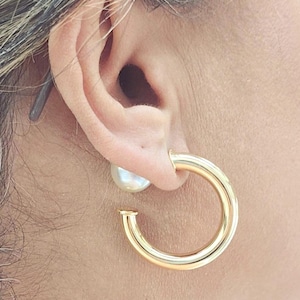 May include: A close-up of a gold hoop earring with a pearl stud. The gold hoop is thick and circular, with a small opening at the bottom. The pearl stud is white and round, positioned behind the earlobe.