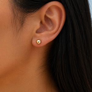 May include: A close-up of a person's ear with a small, round, gold stud earring. The earring is a simple, classic design. The person has dark hair and fair skin. The background is blurred, suggesting a beach setting.