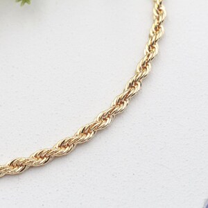 May include: Gold-coloured rope chain necklace. The chain is made of twisted links, creating a rope-like texture. The necklace is displayed on a white surface, with a hint of a light purple background.