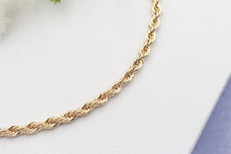 May include: Gold-coloured rope chain necklace. The chain is made of twisted links, creating a rope-like texture. The necklace is displayed on a white surface, with a hint of a light purple background.