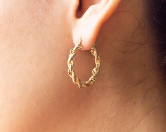 May include: A gold hoop earring featuring a twisted rope texture design. The circular earring is secured through the earlobe, showcasing a polished metallic finish with intricate, repeating diagonal grooves along the entire surface of the hoop.