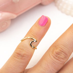 May include: Gold wave ring on a finger. The ring is a simple design with a wave shape.