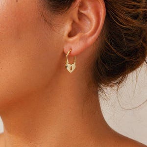 May include: A close-up of a person's ear wearing a gold-colored heart-shaped lock earring. The earring is a hoop style with a heart-shaped charm. The background is blurred, focusing on the earring.