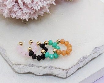 May include: Four beaded rings in pink, black, green, and orange. Each ring is made of small beads with gold accents.