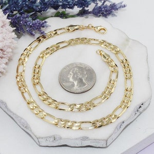 May include: A gold-colored Figaro chain necklace with a lobster clasp, displayed on a white surface. The necklace is next to a quarter coin for size comparison. Decorative flowers are in the background.