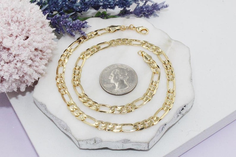 May include: A gold-colored Figaro chain necklace with a lobster clasp, displayed on a white surface. The necklace is next to a quarter coin for size comparison. Decorative flowers are in the background.