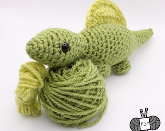 Pode incluir: Um brinquedo de dinossauro de crochê verde feito à mão com uma barbatana amarela, olhos pretos e uma bola de fio verde. O dinossauro está deitado ao lado do fio, em um fundo branco.