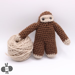 May include: A brown crocheted sloth amigurumi toy holding a ball of beige yarn. The sloth has black eyes and a beige face.