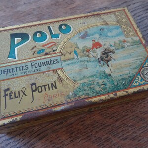 May include: Vintage tin box with a gold background and a blue and white image of a polo match. The text on the box reads "Polo Gaufrettes Fourrees au praline Felix Potin - Paris".