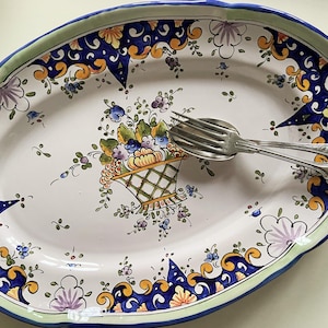 Peut inclure: Un plat de service ovale en céramique blanche avec un motif floral bleu, vert et jaune. Le plat présente un motif floral central avec un panier de fleurs et un bord festonné.