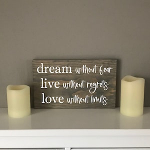 May include: A wooden sign with the text "dream without fear live without regrets love without limits" in white lettering. Two white candles are on a white surface below the sign.