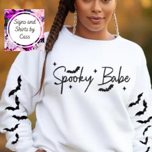 以下が含まれることがあります： 「Spooky Babe」の文字が黒い筆記体で書かれた白いスウェットシャツ。コウモリのシルエットと星で装飾されています。袖にはコウモリのデザインがあります。カジュアルなハロウィーンをテーマにしたアパレルアイテムです。
